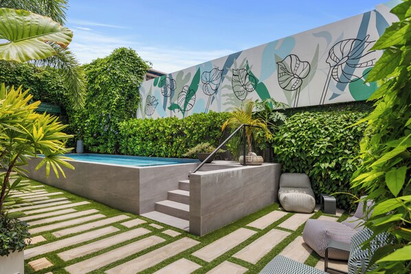 Relax by the pool surrounded by lush greenery and modern furniture, with a pathway leading to the pool area and a vibrant mural adorning the walls.