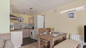 A compact kitchen with a clock, a dining table set with plates and glasses, and a sofa in the living area. - Harbour Reach - Close to Beach Parking (Saundersfoot)