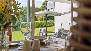A dining table set with plates, glasses, and cutlery, overlooking a garden with a patio chair and umbrella. - Harbour Reach - Close to Beach Parking (Saundersfoot)
