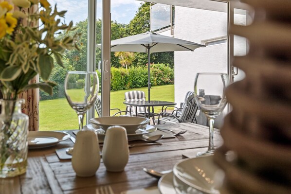 A dining table set with plates, glasses, and cutlery, overlooking a garden with a patio chair and umbrella.