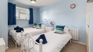 A bedroom with two beds, a window with curtains, a bedside table, a clock, and a radiator. - Harbour Reach - Close to Beach Parking (Saundersfoot)