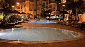 A nighttime pool area with lounge chairs and umbrellas, surrounded by buildings with balconies and palm trees. - Seaview Apartment Estrela da Luz, Praia da Luz (Lagos)