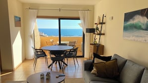A living room with a sofa, a coffee table, and a dining area with chairs and a table. There is a view of the ocean through large windows. - Seaview Apartment Estrela da Luz, Praia da Luz (Lagos)