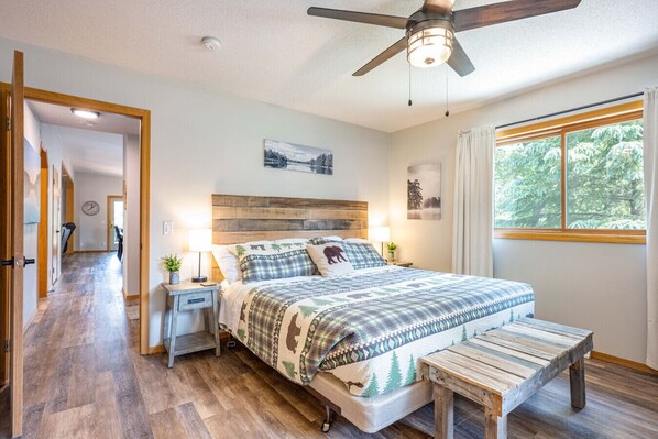A bedroom with a wooden headboard, a bed with a patterned blanket, bedside tables with lamps, a bench, and a large window with a view of trees. - Paddler`s Paradise at Lake McGinnis (Oxford)