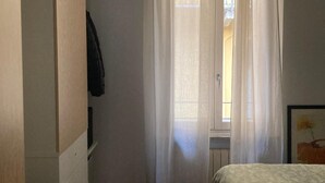 A small, well-lit bedroom with a bed, a wardrobe, and a window with sheer curtains.