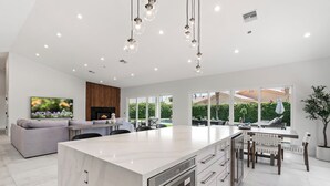 A modern kitchen with a large island, built-in oven, and stainless steel appliances. There is a dining area with a table and chairs, and a living room with a sofa and TV.