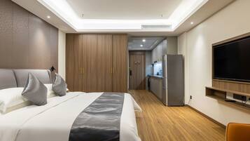 A modern hotel room with a large bed, a TV mounted on the wall, a kitchen area, and wooden flooring.