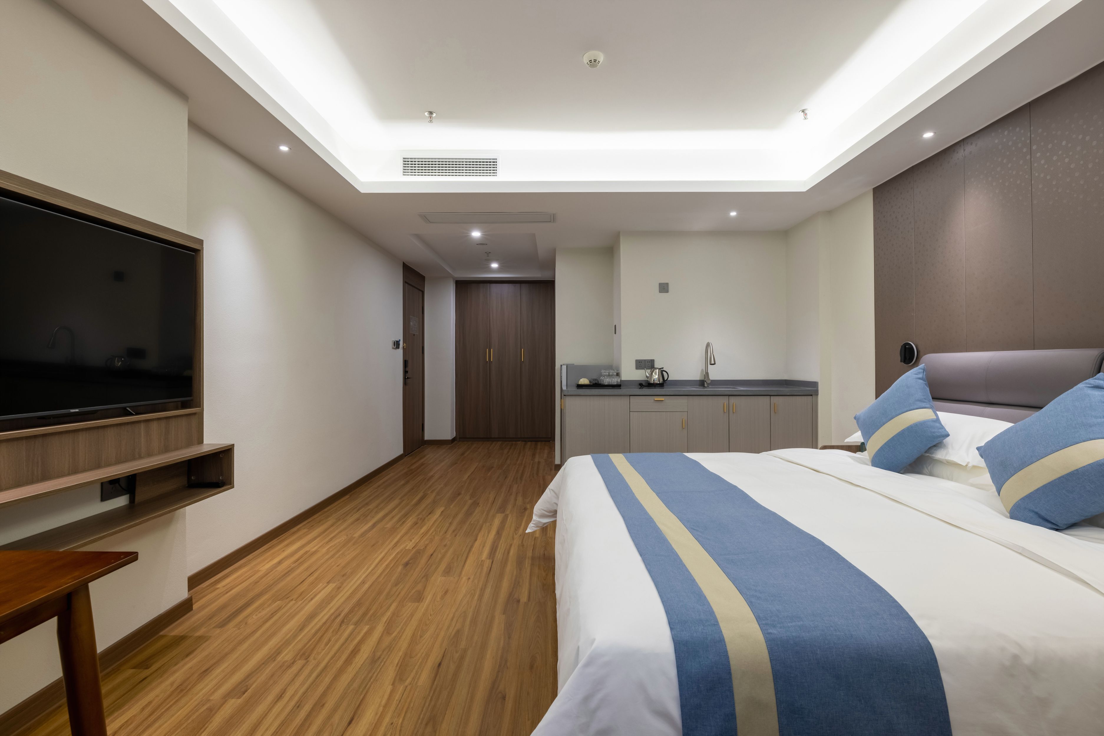 A modern hotel room with a large bed, a TV mounted on the wall, a small kitchenette, and a wooden floor.