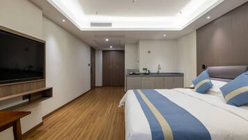 A modern hotel room with a large bed, a TV mounted on the wall, a small kitchenette, and a wooden floor.