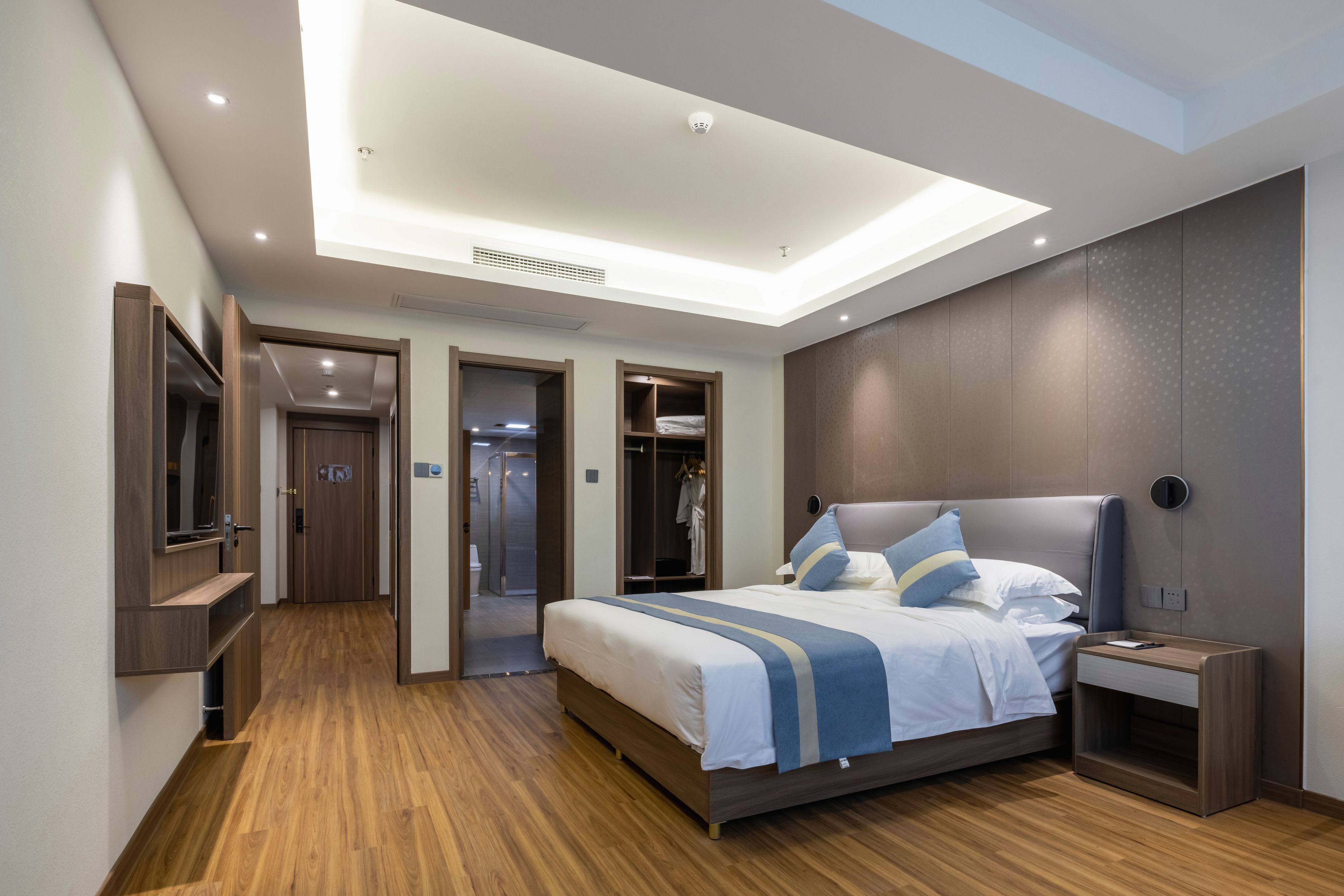 Ein modernes Hotelzimmer mit einem großen Bett, einem Nachttisch, einem integrierten Fernseher und einem begehbaren Kleiderschrank.