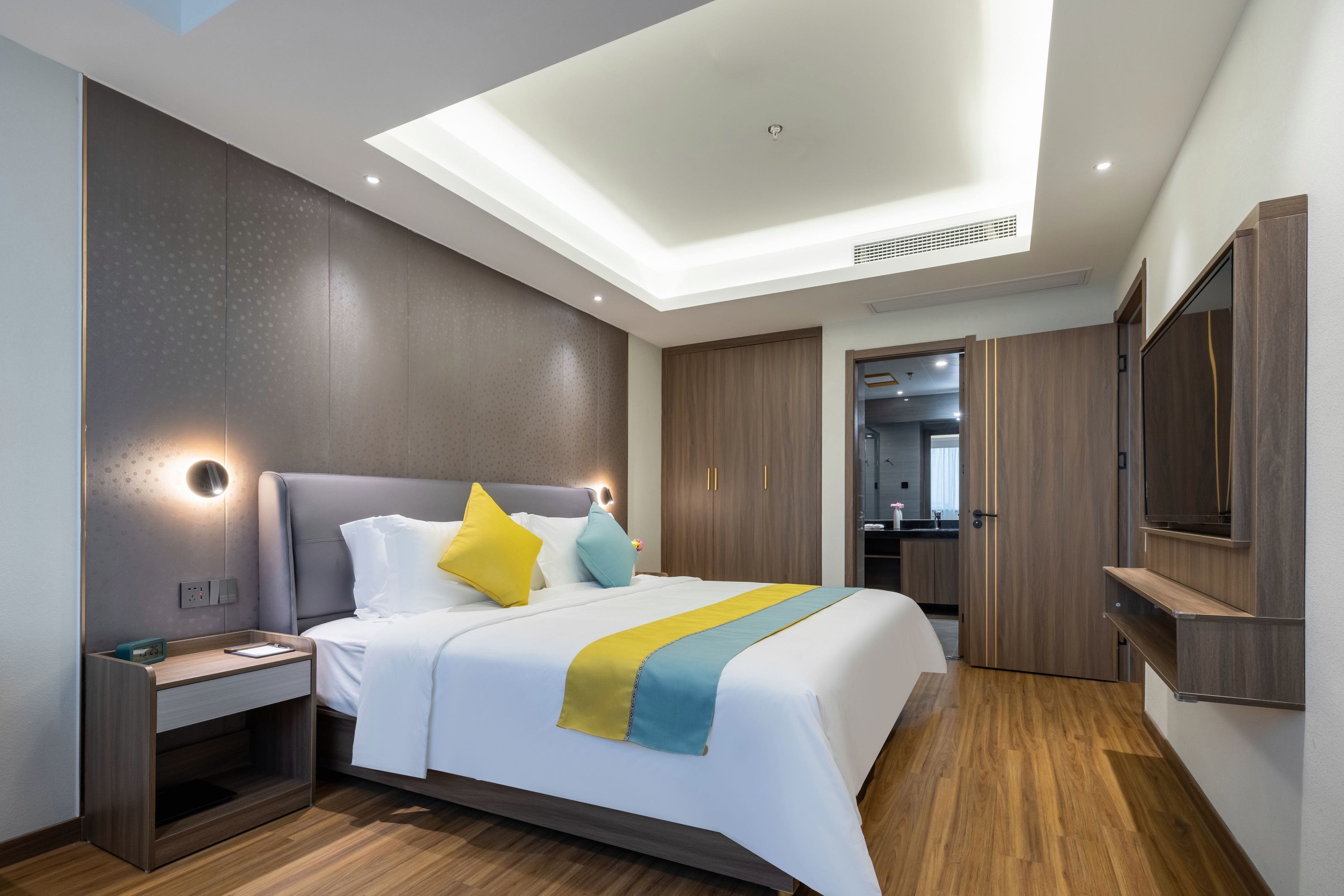 A modern hotel room with a large bed, built-in nightstand, and a flat-screen TV mounted on the wall.