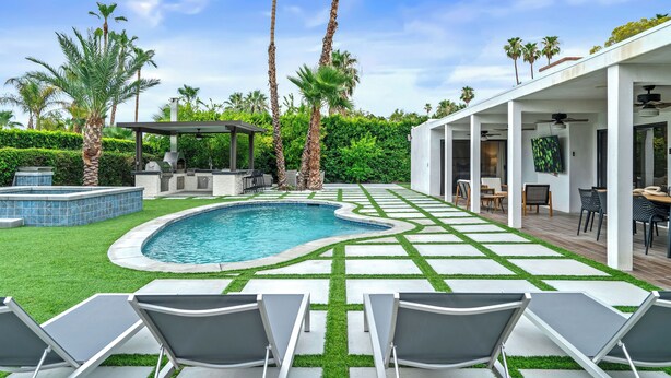 A backyard with a swimming pool, lounge chairs, palm trees, and a barbecue area.