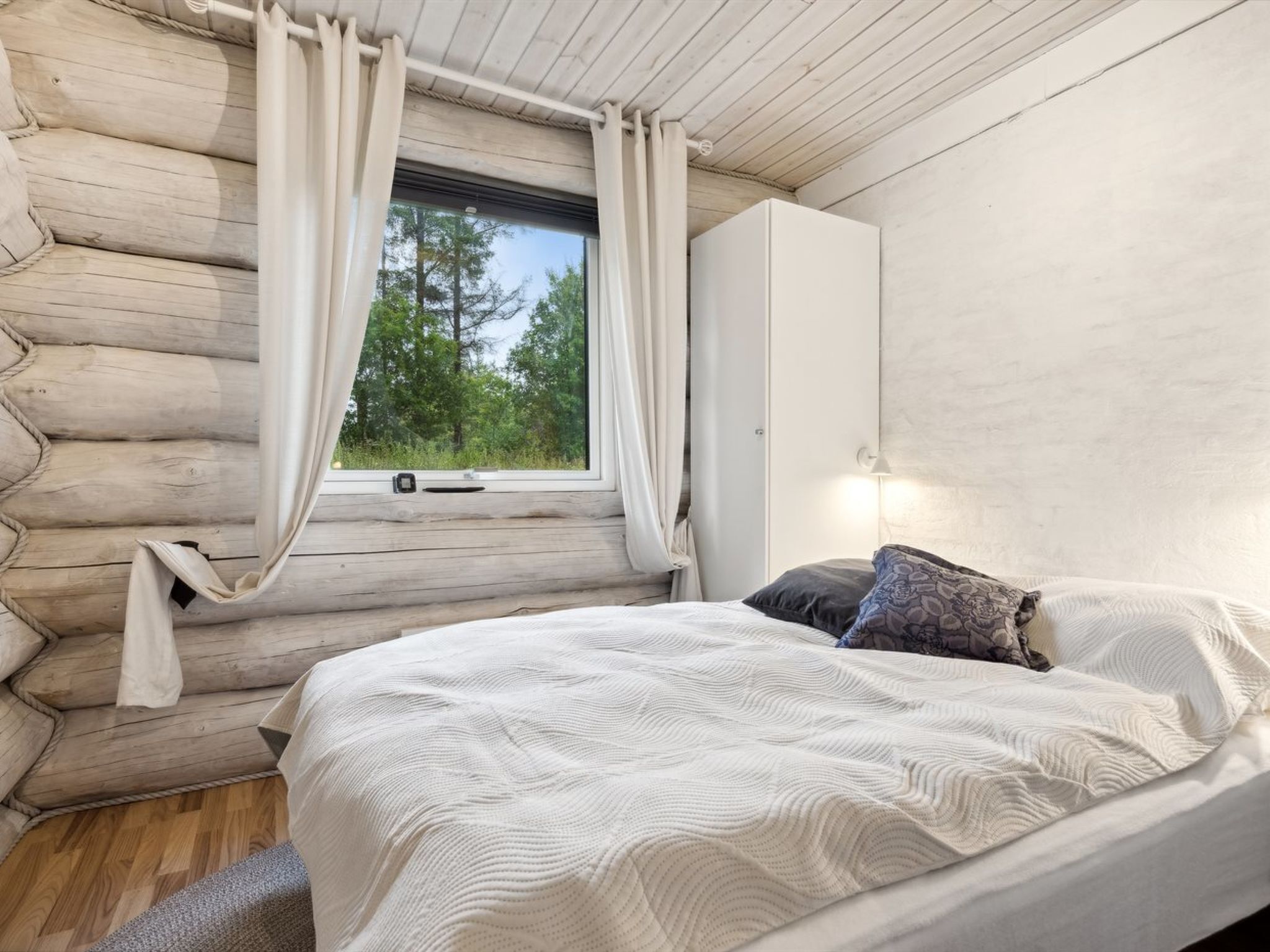 Ein Schlafzimmer in einer Blockhütte mit einem Bett, einem Fenster mit Vorhängen und Blick auf Bäume.