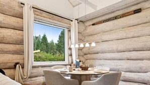 Een blokhut van binnen met een eettafel, stoelen, een lamp en uitzicht op groen buiten.