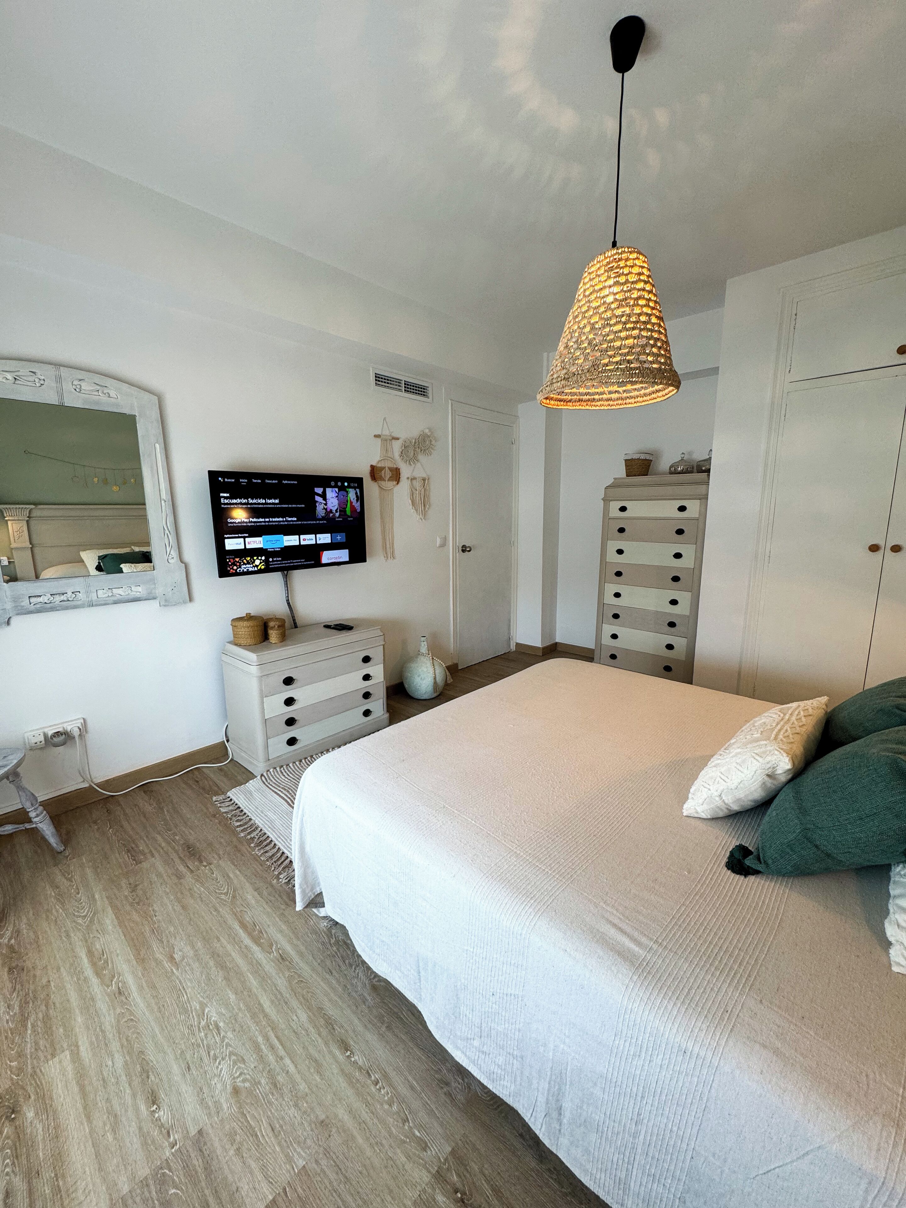 A bedroom with a large bed, a dresser, a vanity with a mirror, a television on the wall, and a hanging light fixture.