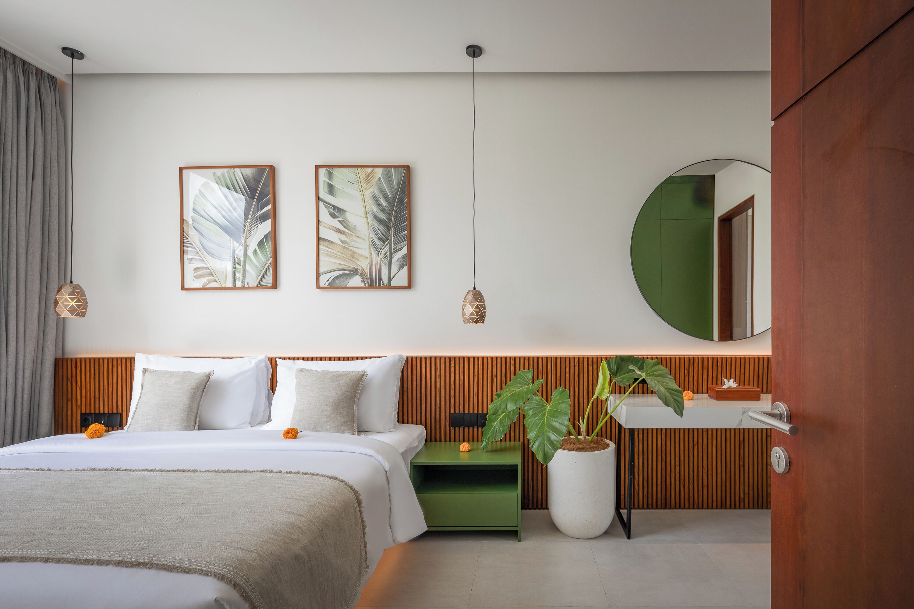 Une chambre d’hôtel moderne avec un lit, une table de chevet verte, une plante en pot, un miroir et une œuvre d’art accrochée au mur.