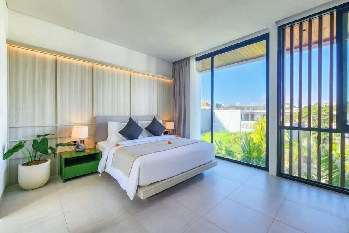 A modern bedroom with a large bed, a nightstand, a potted plant, and a view of greenery through large windows.