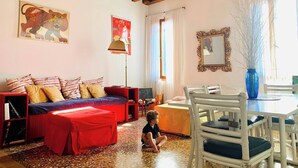 A cozy living room with a sofa, a child playing, a dining table with chairs, a vase with branches, and artwork on the walls. - Painters House Biennale Arsenale (Venice)
