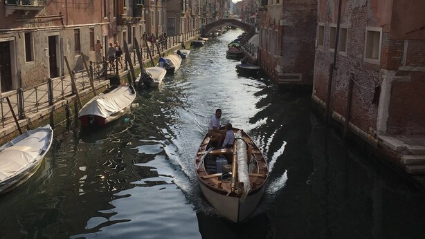 A narrow canal with boats, buildings on either side, and a bridge in the distance. - Painters House Biennale Arsenale (Venice)