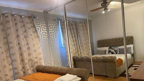 Un dormitorio moderno con un espejo grande, una cama con una manta naranja y una segunda cama con cabecera capitonada.