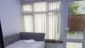 Habitación compacta con sofá cama gris, un radiador, una ventana con cortinas y vistas a una terraza de madera.