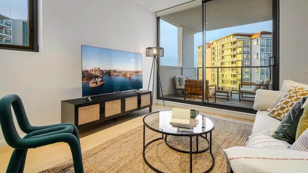 A modern living room with a large TV, comfortable seating, and a balcony with a city view.