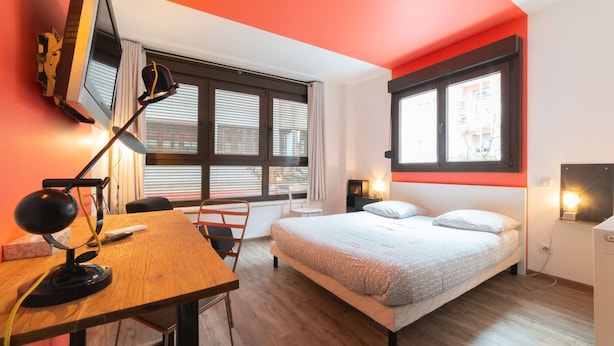 A modern bedroom with a wooden desk, a bed with patterned bedding, and a large window with blinds. - Chambre privative rouge (Mulhouse)