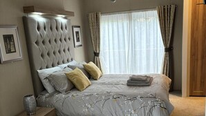 Une chambre à coucher avec une tête de lit capitonnée, une grande fenêtre ornée de rideaux légers, une table de chevet avec un vase et un lit recouvert d’une literie à motifs floraux.