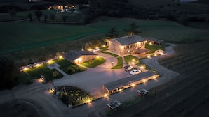 Luchtbeeld van een stenen huis met een oprit, een overdekt terras en een verlicht landschap in de schemering.