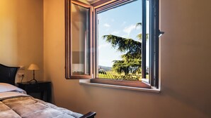 Een slaapkamer met een bed, een nachtkastje, een lamp en een raam met uitzicht op een wijngaard en bomen.