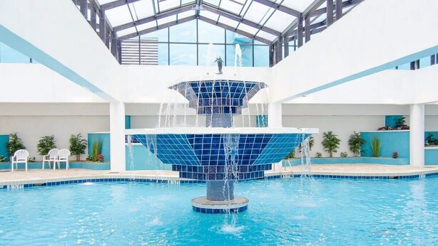 Une grande piscine intérieure dotée d’une fontaine centrale, entourée de chaises blanches et de plantes en pot.