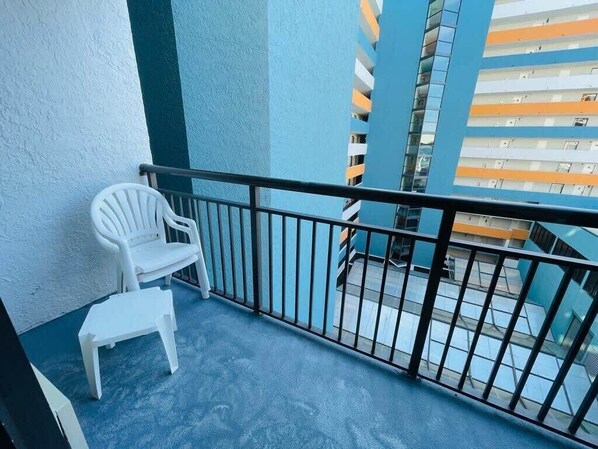 A balcony with a white plastic chair and a small table, overlooking a building with multiple balconies. - LA82 Beautiful Studio on the beach + pool+ hot tub (Myrtle Beach)