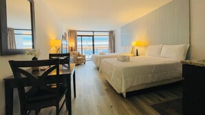 Habitación de hotel con dos camas, un escritorio, una silla y un amplio ventanal con vistas al mar.