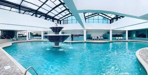 Une piscine intérieure dotée d’une fontaine centrale, entourée de chaises longues et surmontée d’un toit en verre.