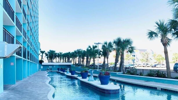 A swimming pool with blue-tiled edges, surrounded by palm trees and a building with blue columns. - LA12 Wonderful condo on the beach + pool + Hot tub (Myrtle Beach)
