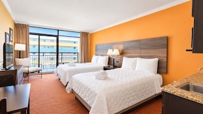 A hotel room with two beds, a desk, a chair, a balcony with a view, and a kitchen area.