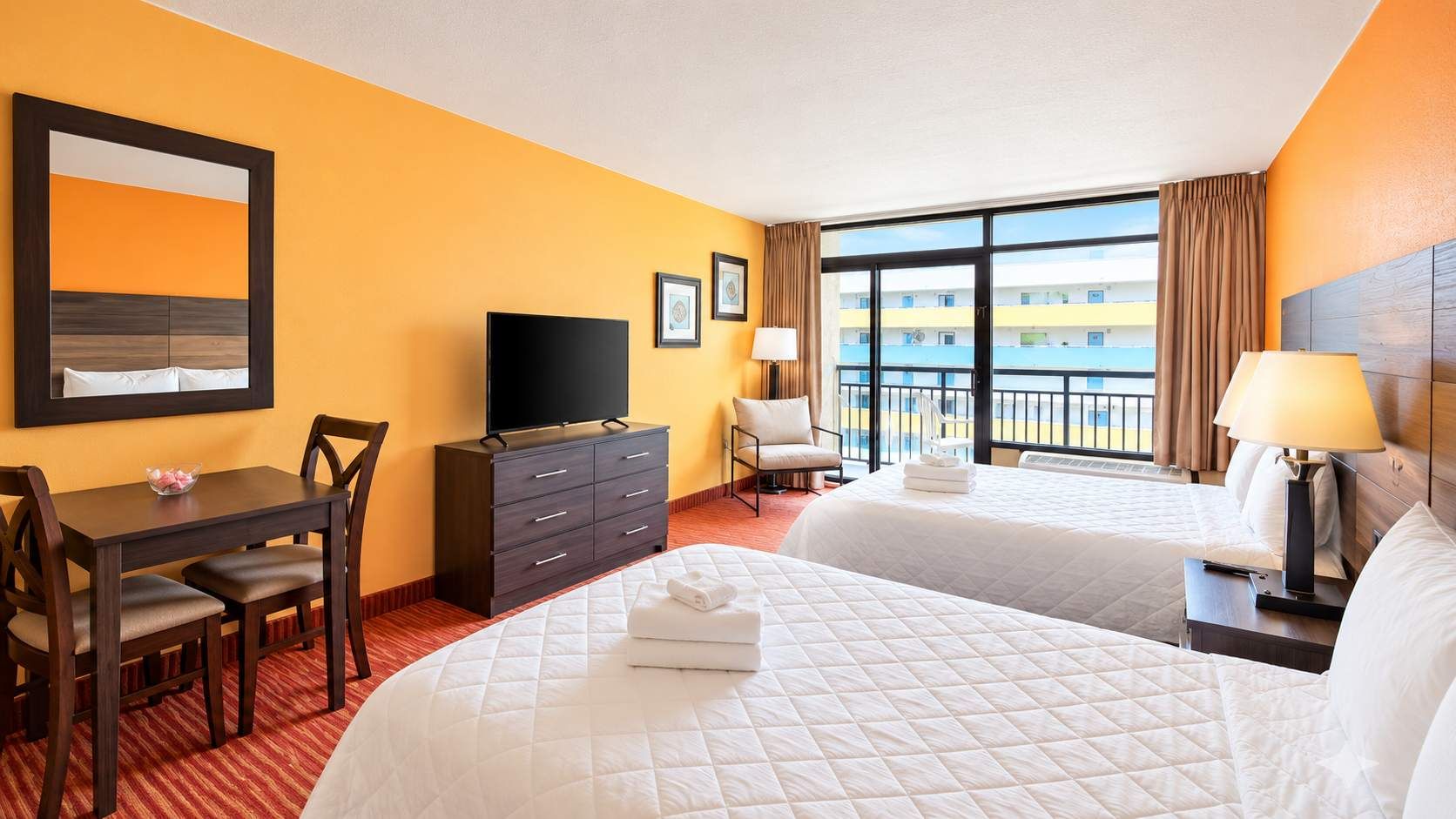 A hotel room with two beds, a desk, a TV, and a view of a building.