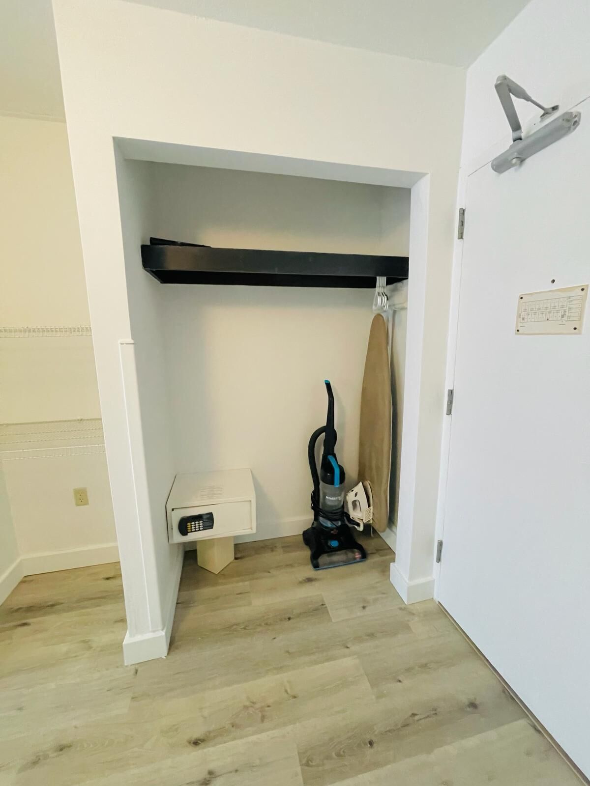 A room with a white door, a vacuum cleaner, a small white cabinet, and a wooden shelf.