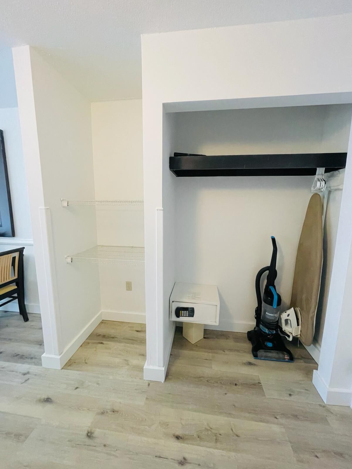 A room with a white wall, a black shelf, a white bench, and a vacuum cleaner.