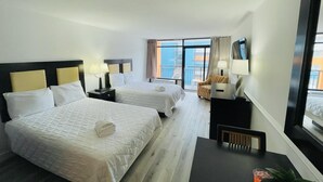 Ein Hotelzimmer mit zwei Betten, einem Schreibtisch, einem Sessel, einem Fernseher und einem Fenster mit Blick auf Gebäude.