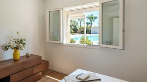 A bedroom with a bed, a bedside table with a vase of flowers, and a window with a view of a pool and greenery.