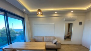 A modern living room with a sofa, wooden dining table, and chairs. There is a large window with a view of the sea and a balcony. - Atlas homes (Demre)