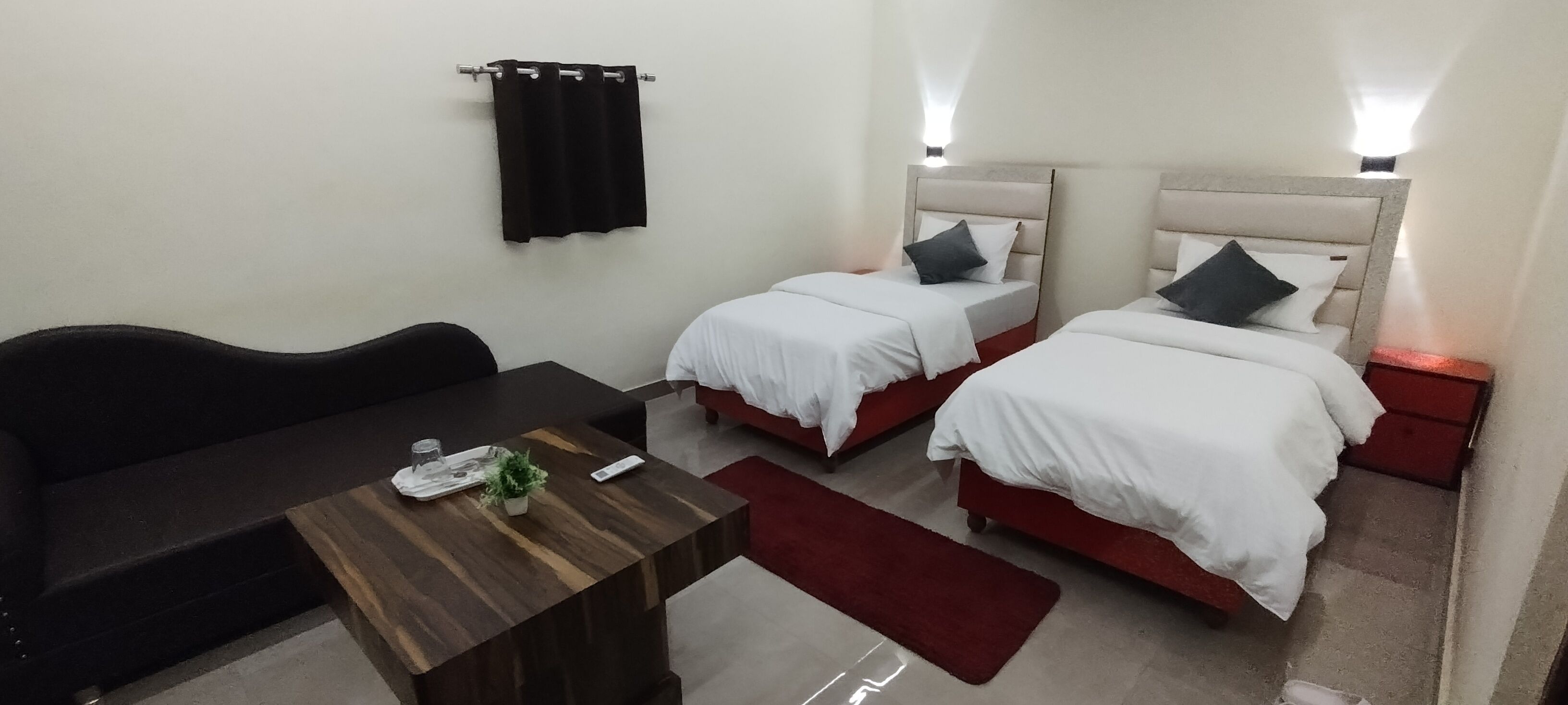 A hotel room with two single beds, a wooden table with a tray and remote, a red rug, and a black sofa.