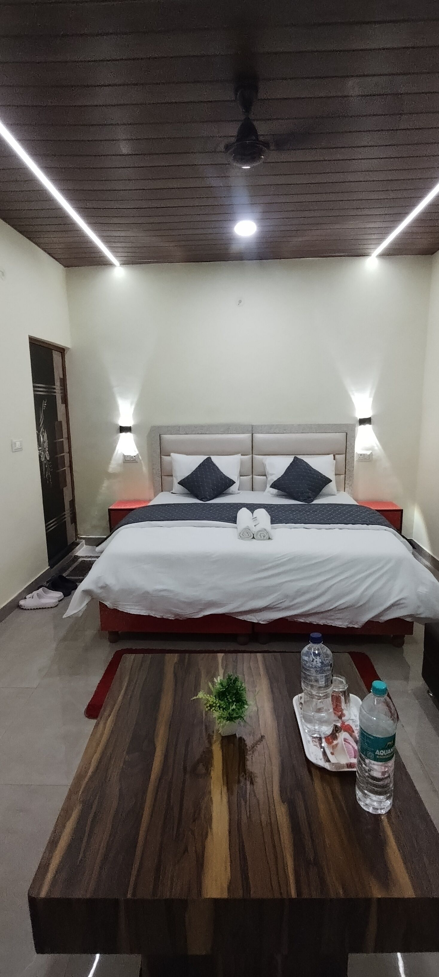 A neatly arranged bedroom with a bed, bedside tables, a wooden table with a plant, and a water bottle.