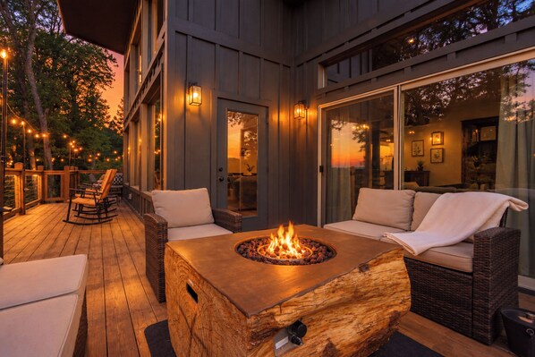 An outdoor deck with a fire pit, wicker furniture, and a view of the interior through large glass doors.