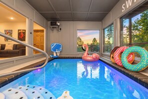 An indoor swimming pool with a diving board, inflatable toys, and a view of the outdoors through large windows.