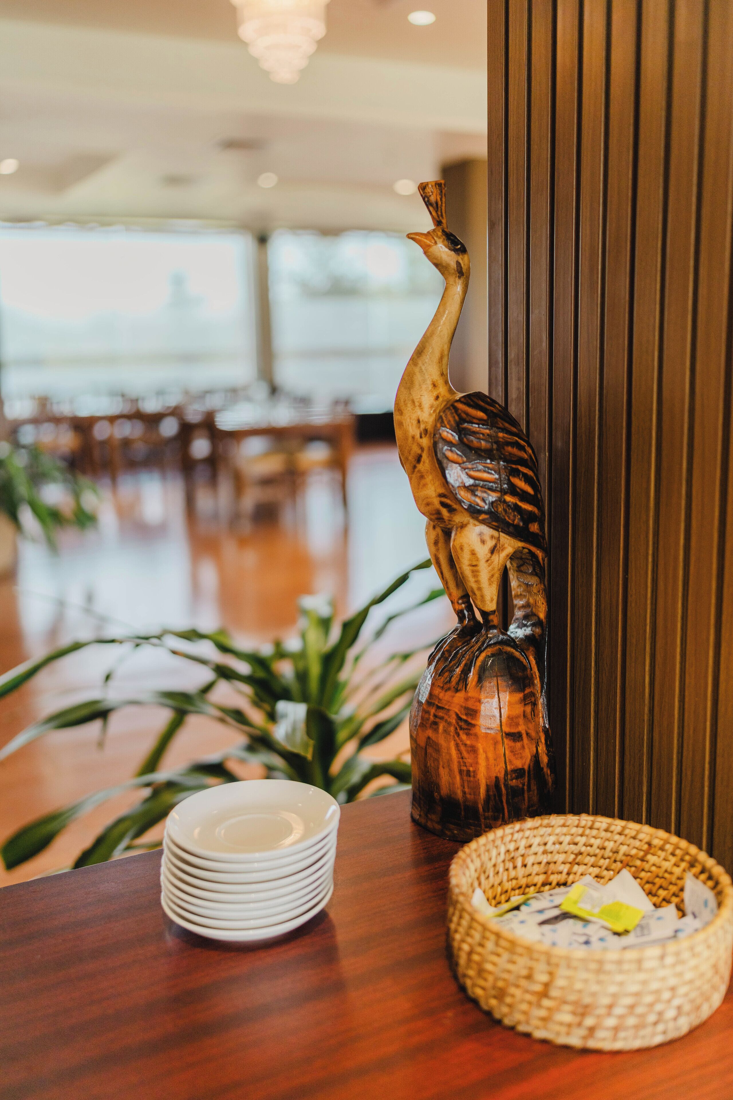 A wooden sculpture on a table, a stack of white plates, and a woven basket with papers.