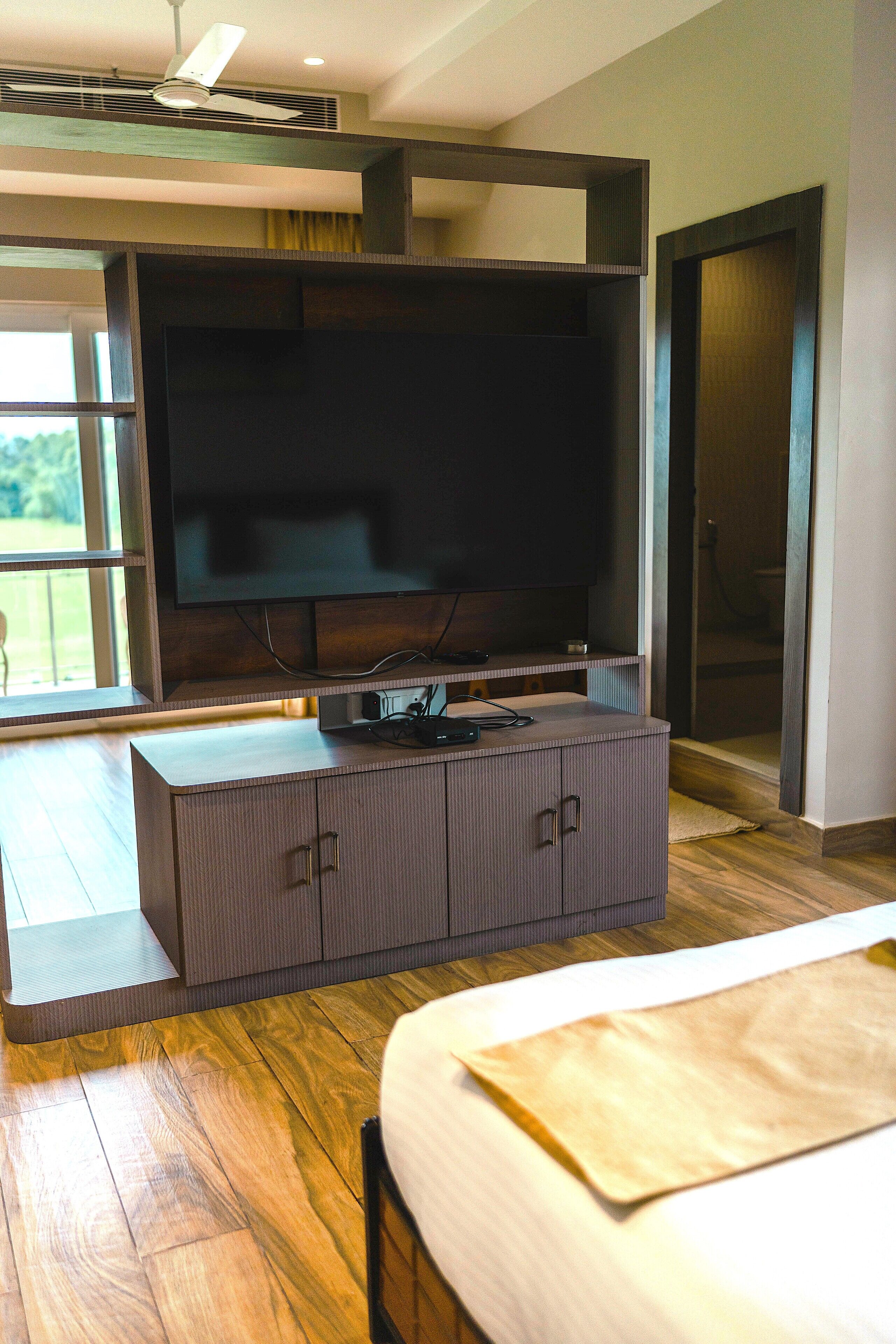 A modern bedroom with a large flat-screen TV mounted on a wooden cabinet, a bed with a white mattress, and a wooden floor.