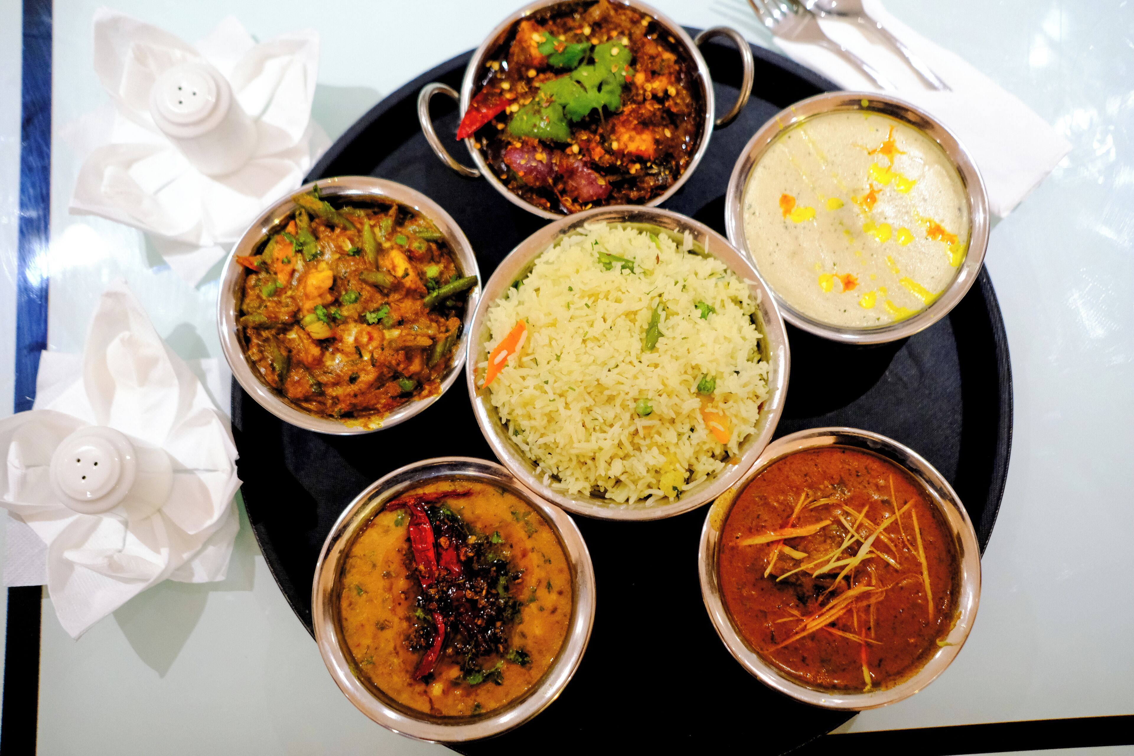 A variety of dishes including rice, curries, and a soup, served on a black tray with white napkins.