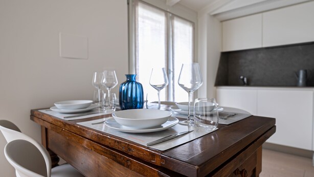A dining table set with plates, bowls, and wine glasses, with a blue vase and a kitchen in the background. - Volta 40 - A in Monza (Monza)